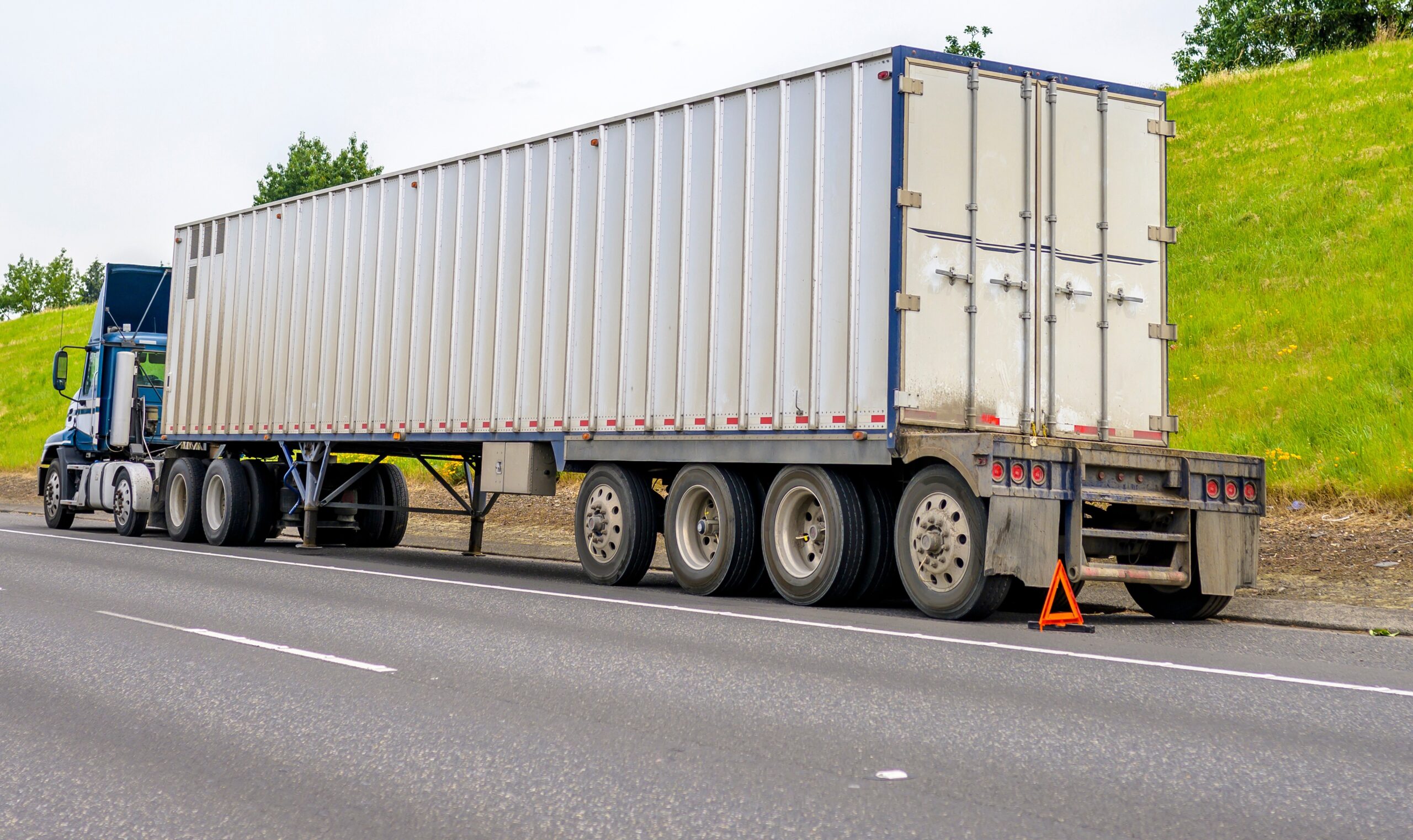 Big rig blue semi truck with loaded container on semi trailer standing broken on the road shoulder set an emergency triangle stop sign behind - RapidLink Repairs™ Blue semi-truck with a long silver trailer stopped on the roadside; a safety triangle sits on the pavement behind it with a grassy hill in the background.