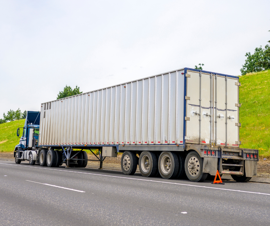 Big rig blue semi truck with loaded container on semi trailer standing broken on the road shoulder set an emergency triangle stop sign behind